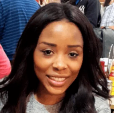 Smiling woman with long black hair wearing a gray top in an indoor setting with people in the background.