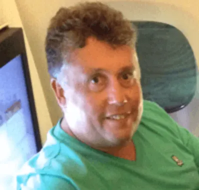 Man with short curly hair wearing a green shirt, seated inside an airplane near a window.