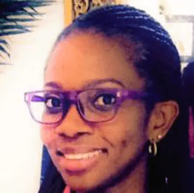 Close-up of a smiling woman with braided hair, wearing purple glasses and gold hoop earrings.