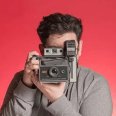 Person with dark hair wearing a gray sweatshirt holding a vintage camera up to their face against a red background.
