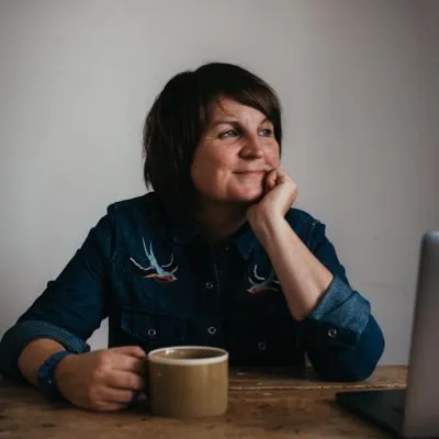 Woman in a denim shirt sitting at a wooden table holding a coffee mug and looking thoughtfully to the side.