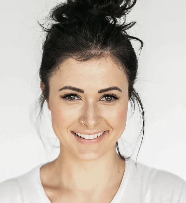 Smiling woman with dark hair in a messy bun, wearing a white shirt and a nose ring, against a plain light background.