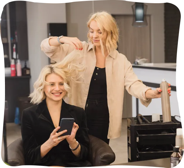 Blonde hairdresser styling a smiling blonde woman’s hair in a salon while the woman looks at her phone.