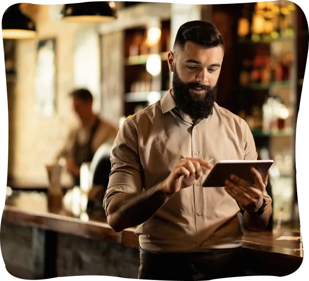 Bearded man in a restaurant using a tablet, standing by the bar counter with shelves of bottles in the background.