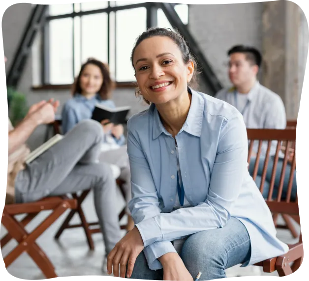 Smiling woman in light blue shirt sitting and leaning forward in a casual group discussion with three people in the background.