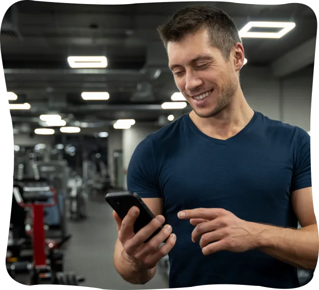 Smiling man in navy blue t-shirt looking at his smartphone in a gym with exercise machines in the background.
