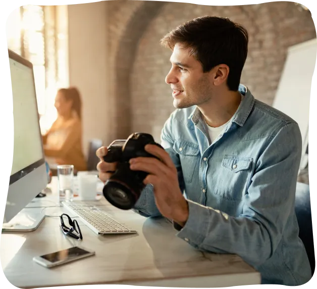 Young man holding a camera and smiling while sitting at a desk with a computer, smartphone, and glasses in a bright office.