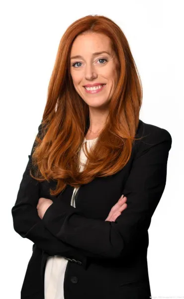 Smiling woman with long red hair in a black blazer and white top, arms crossed against a white background.